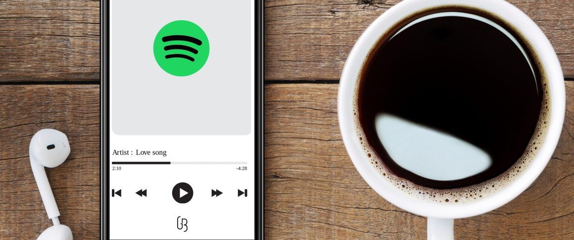 Foto de um celular e uma xícara de café em uma mesa de madeira. Na tela do celular, aparece uma música tocando, a marca do Spotify e da marca Guilherme Barbosa.
